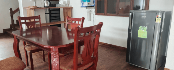 A red wood dining table set with six matching chairs in a kitchen area, black refrigerator and cabinets visible in the background.