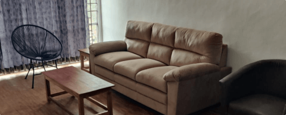 A simple living room interior with brown leather sofa, wooden coffee table, and armchair. The room has beige walls, hardwood floors, and curtained windows.