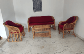 A room with a wicker furniture set comprising a sofa, two armchairs with red cushions, and a coffee table, all placed on a marble floor. A small window with a curtain is visible in the background.