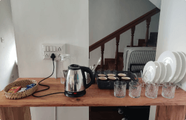 Kitchen counter setup with an electric kettle, a basket of tea and coffee packets, six glasses, and a stack of plates next to a staircase