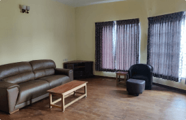 A simple living room interior with brown leather sofa, wooden coffee table, and armchair. The room has beige walls, hardwood floors, and curtained windows.