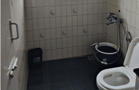 Bathroom with tiled walls, a white toilet, a small black trash can, and a bucket with a faucet and showerhead attached to the wall.